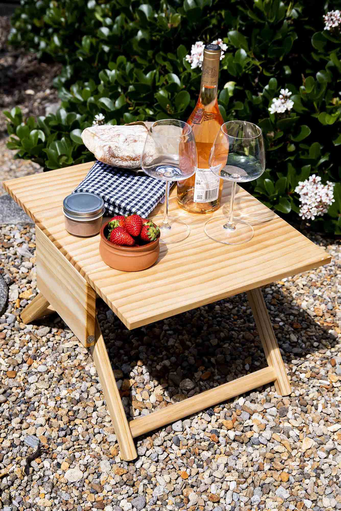 Small wooden table with a bottle of wine, glasses, and snacks on a gravel surface with greenery in the background. Victoria James Home Storage Box VJA Convertible