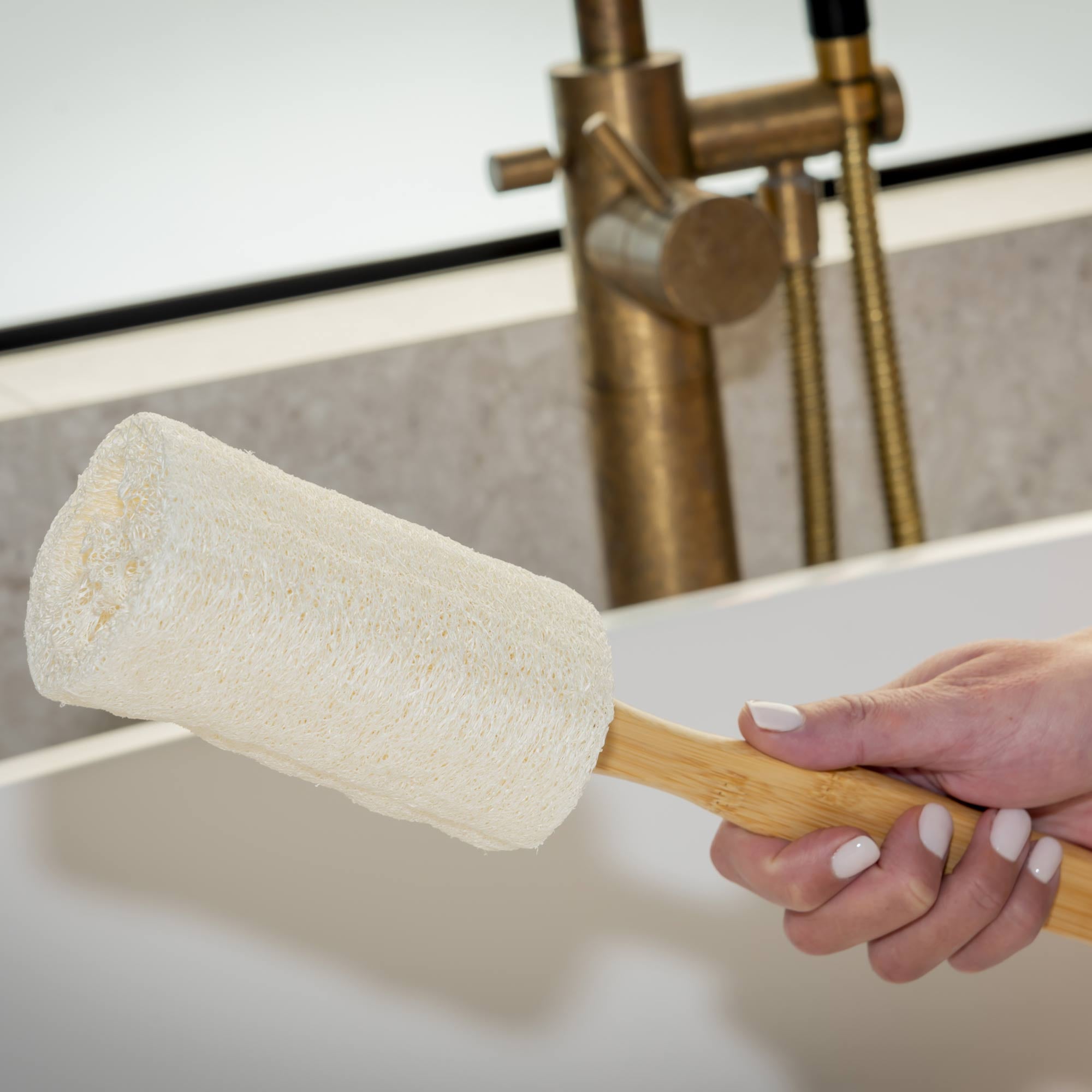 Person holding a wooden scrubber with a white sponge over a sink in a bathroom setting. Victoria James Body Loofah Brush