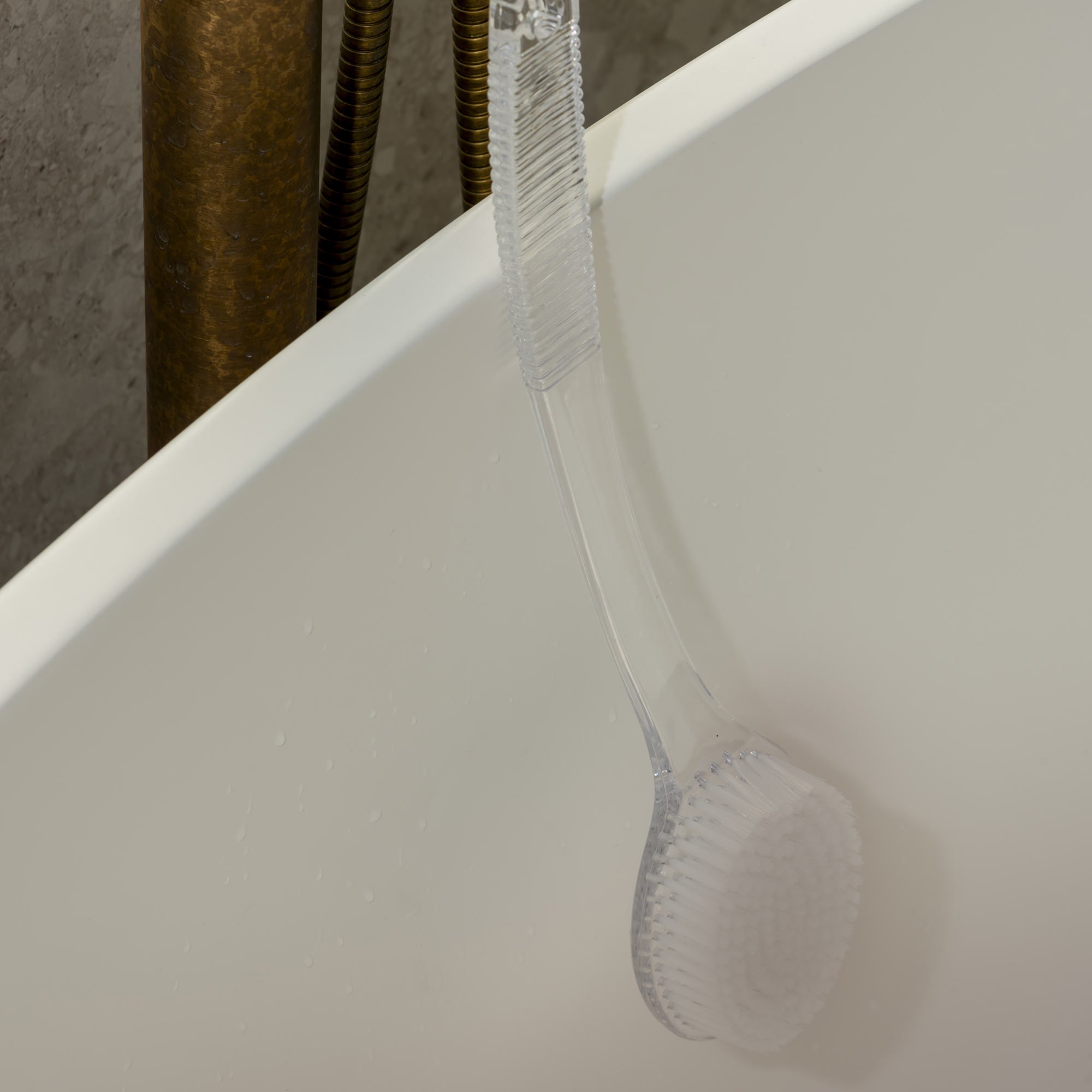 Clear shower curtain liner on a white bathtub with a metallic shower head in the background. Victoria James Body Bath & Shower Brush
