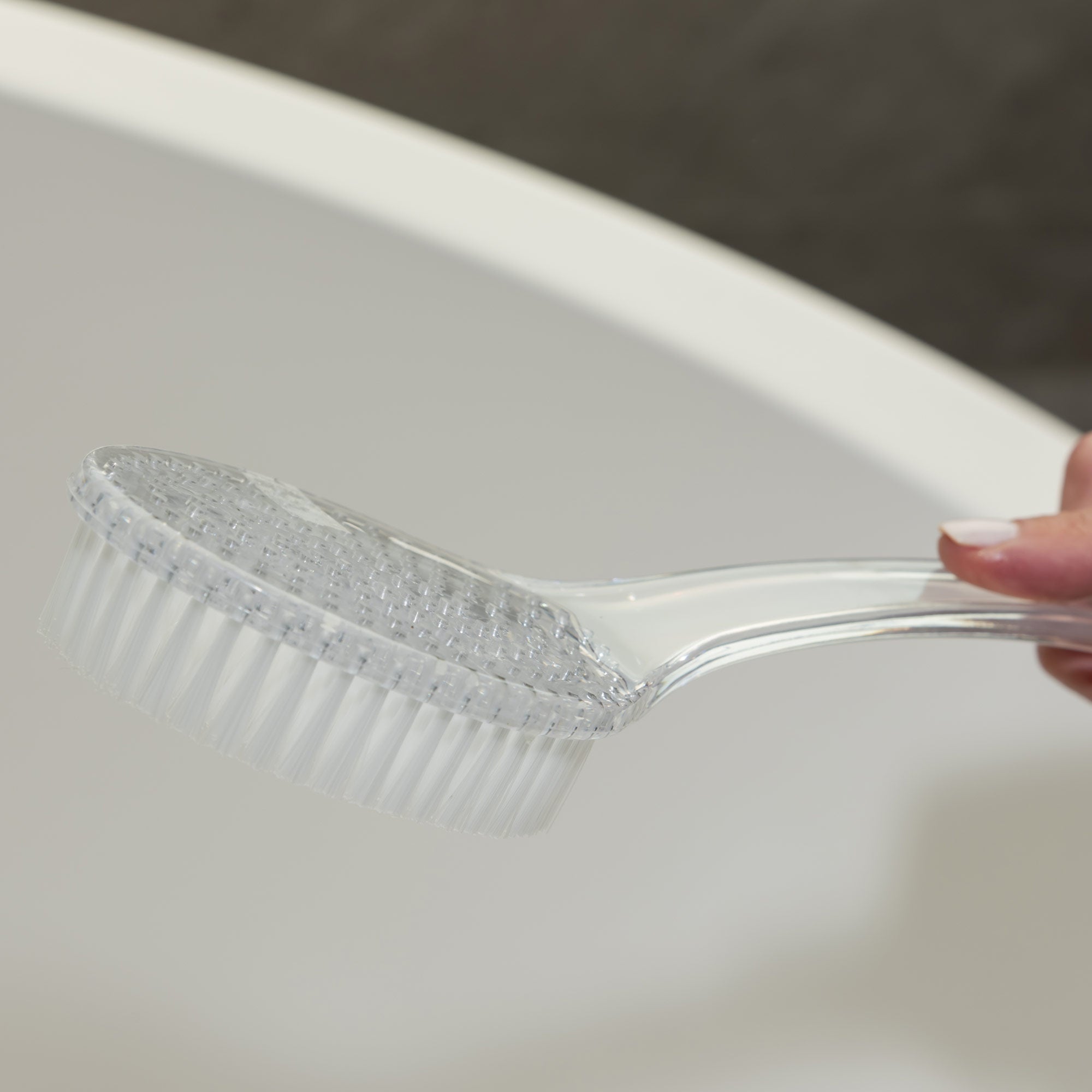 Clear plastic scoop held above a white container with a neutral background. Victoria James Body Bath & Shower Brush