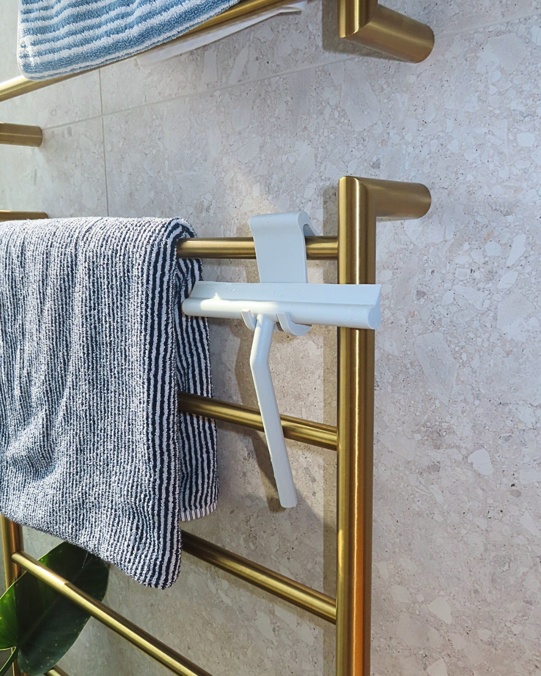 Gold towel rack with a white hook and gray towel on a marble wall. Simply Clean Squeegee