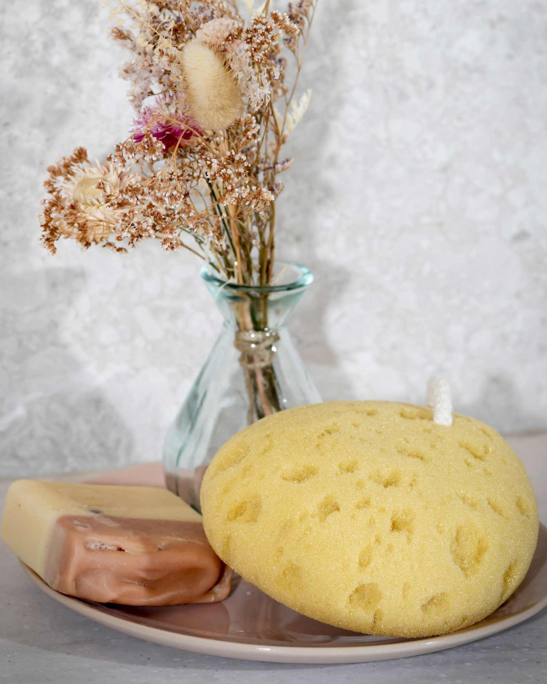 Yellow sponge, bar of soap, and dried flowers in a glass vase on a white surface. Simply Clean Victoria James Body Sponge