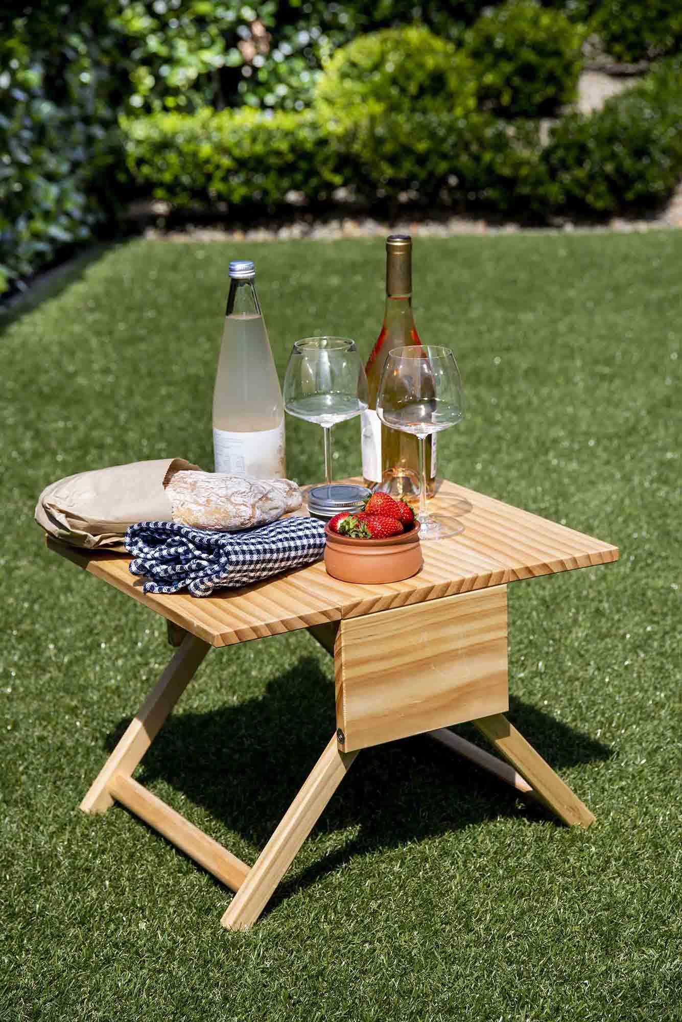Wooden outdoor table with bottles, glasses, and snacks on a grassy background. Victoria James Home Storage Box VJA Convertible